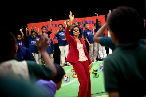 First Lady Michelle Obama in a r4ed suit dancing with her eyes close with a group of girls in blue shirts.