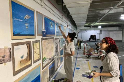 Yvette Dibos stretches her paintbrush to a bird on a long mural with various framed birds, while Laura Sanchez stands back and looks at another bird painting.