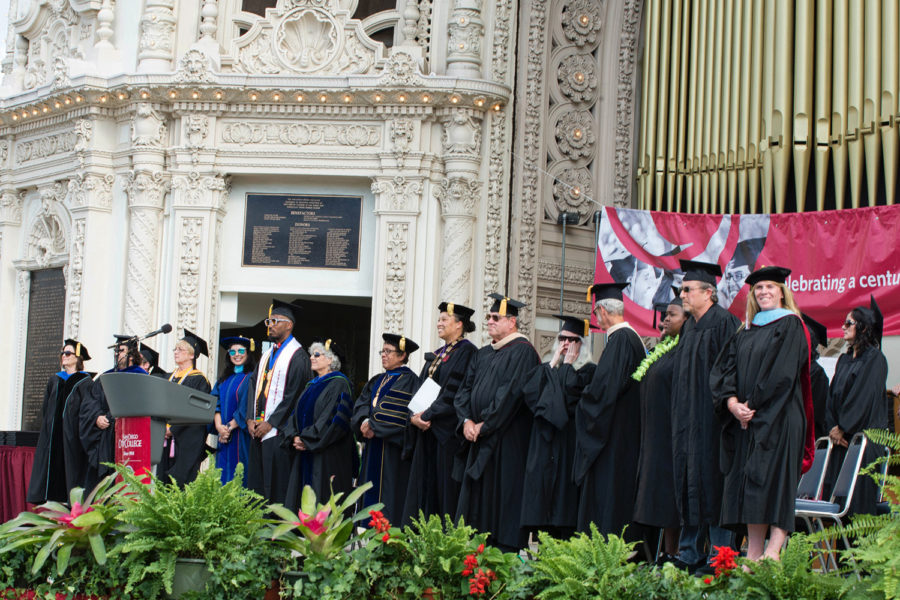 Graduates from City College in caps and gowns stand in a row at the Organ Pavilion in Balboa Park.