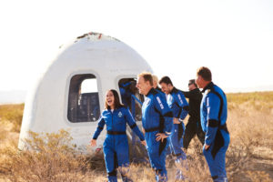 Six astronauts in blue suits stand on the ground outside of the Blue Origin New Shepherd capsule.