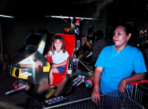 Katya Echazarreta sits in an arcade helicopter with a red seat and her mother, Lilian Martin, stands next to her in a blue shirt.