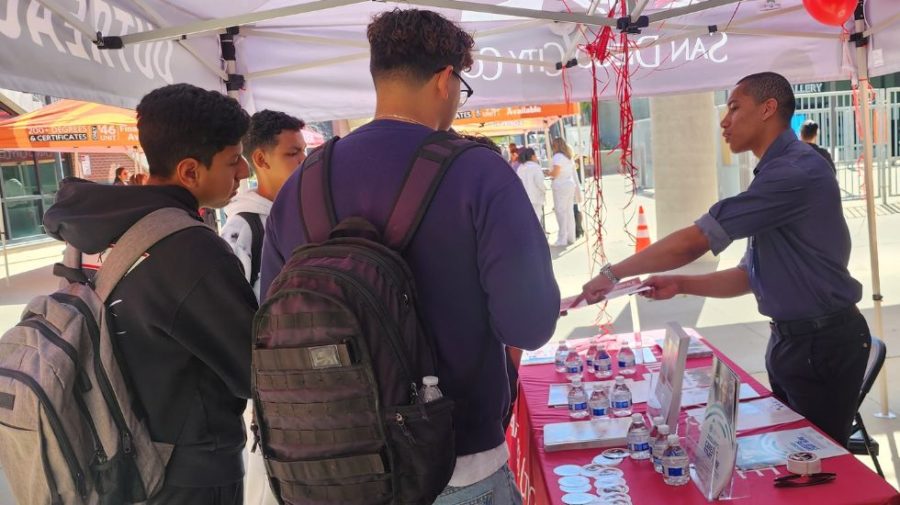 San Diego City College ASG president Diego Bethea stands behind a table and hands out items to three students standing in front of the table.