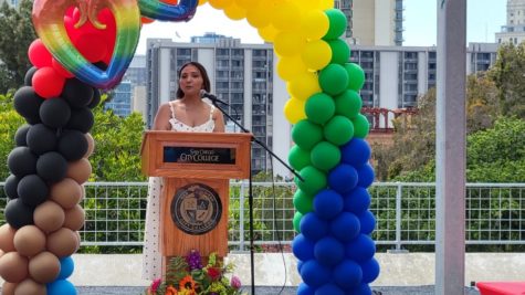 Lucy Plascencia stands in front of a wooden podium surrounded by an arch of yellow, green and blue balloons on an outdoor patio with a building in the background.