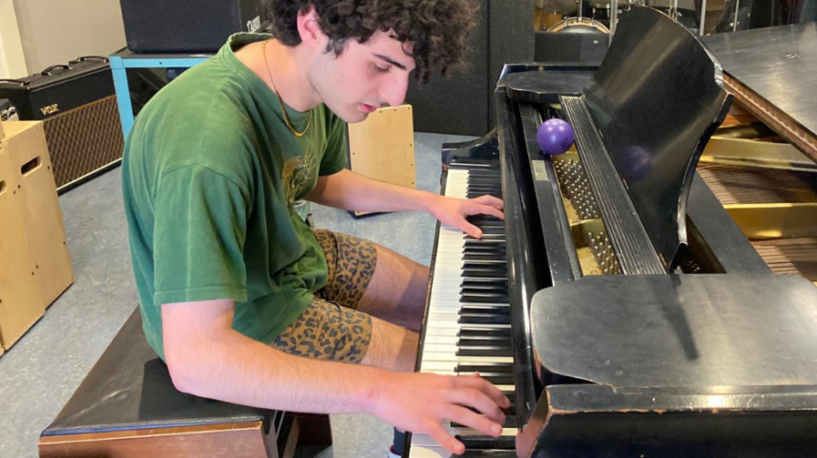 A young man in a green t-shirt plays a black piano in a cluttered recording studio.