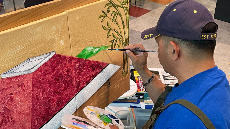 A man in a blue shirt and blue hat uses a fine tip brush to add green paint to a large tan canvas. April 25, 2023. Photo by Kathryn Gray/City Times Media