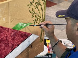 A man in a blue shirt and blue hat uses a fine tip brush to add green paint to a tan canvas
