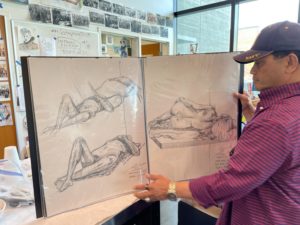 A man in a blue hat and a red and blue striped button down shirt holds open a large art portfolio and displays two pages of drawings.