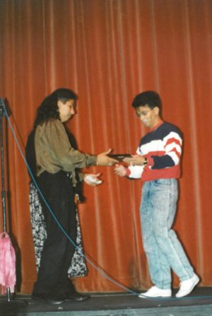 A man hands another man a small plaque while standing on a black stage with a red curtain in the background