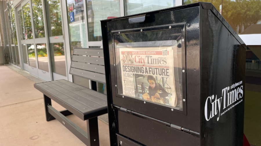 A black newspaper stand displays a newspaper in front of a building and next to a bench.