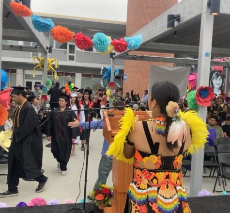 Professor stands at podium dressed in traditional Aztec garb as graduates walk down the aisle