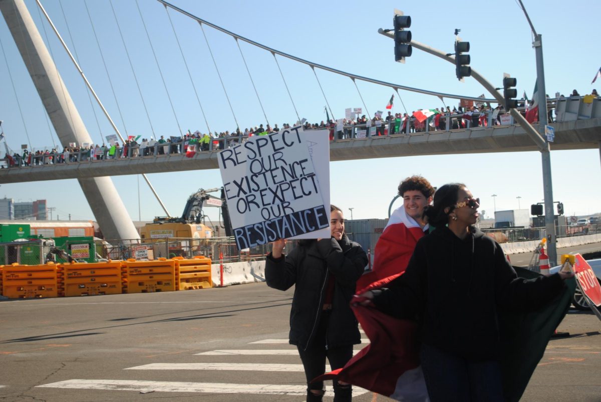 Three demonstrators cross Park Boulevard towards a growing crowd Sunday, Feb. 02, 2025 while holding a sign that says, “Respect Our Existence or Expect Our Resistance.” Photo by Keila Menjivar Zamora/City Times Media