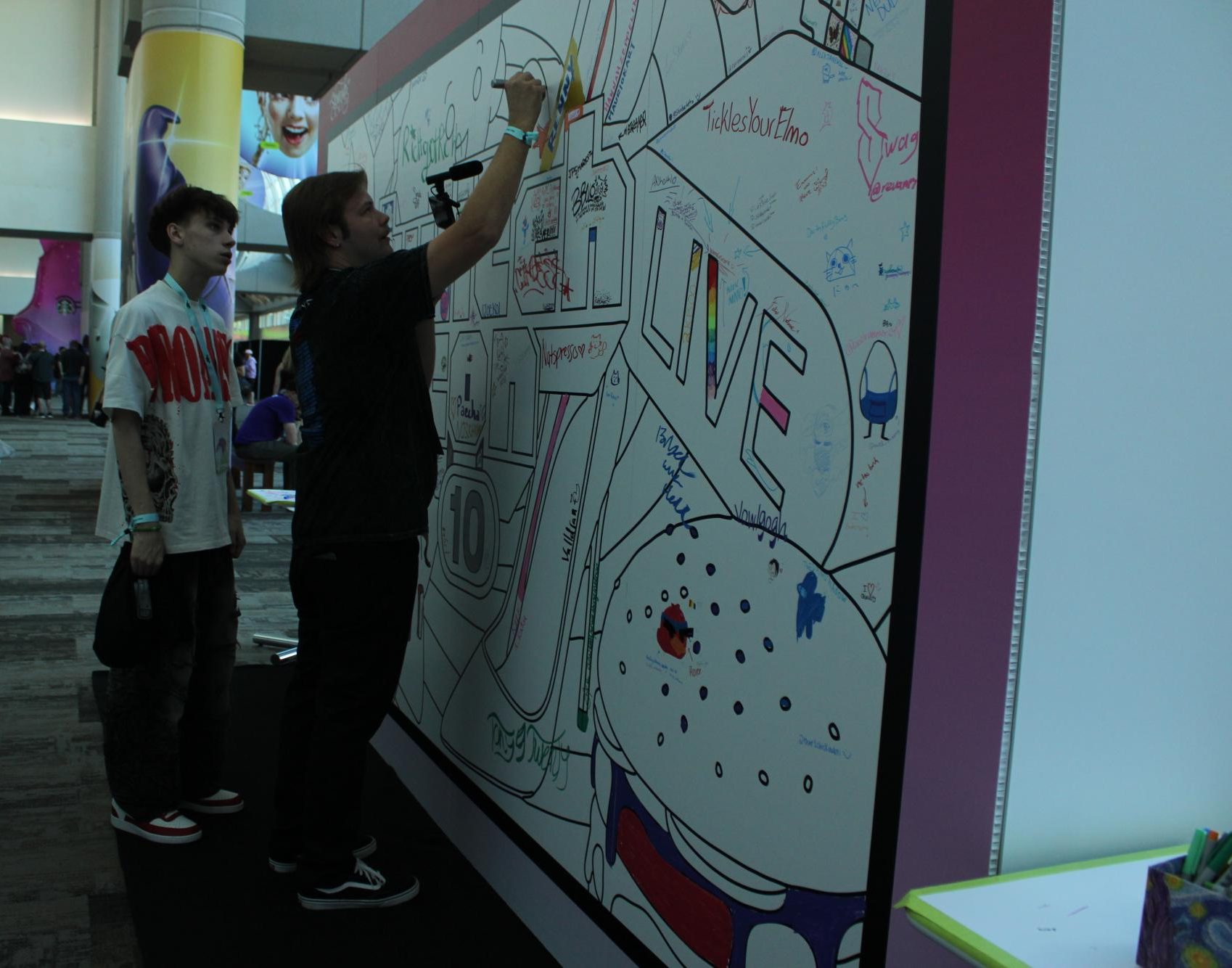 Streamers Humidhimself, left, and Topnotchdrew, right, sign their names on the wall at the doodle hub at the San Diego Convention Center, Friday, Oct. 18, 2025. Photo by Tresean Osgood/City Times Media